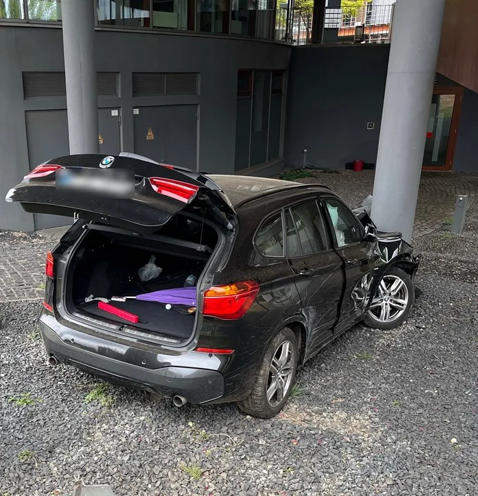 Ein schwarzer BMW zeigt erhebliche Frontschäden und einen geöffneten Kofferraum. Das Fahrzeug steht neben einem Gebäude auf Kies.