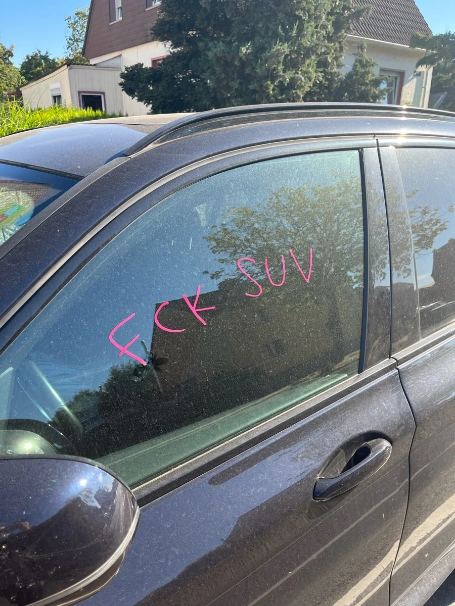 Ein schwarzer SUV steht am Straßenrand einer Wohngegend. Auf der Seitenscheibe ist ein Graffiti mit pinker Farbe.