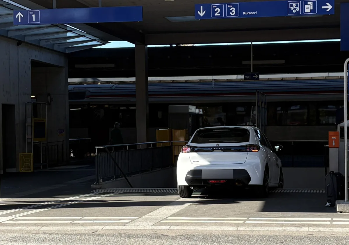 Ein weißes Auto parkt vor einem Eingang zu den Gleisen 2 und 3 am Bahnhof Kurzdorf. Im Hintergrund sind Zugwagen sichtbar.