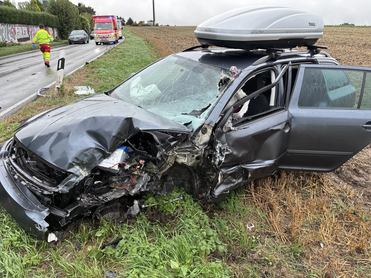 Ein graues Auto mit schweren Frontschäden steht am Straßenrand. Ein Rettungswagen ist im Hintergrund auf der Straße sichtbar.