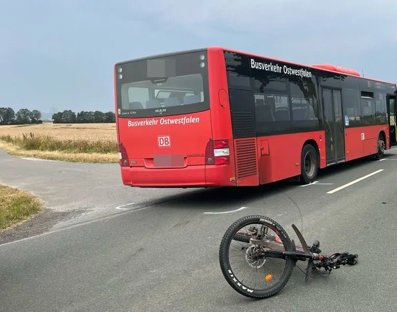 Ein roter Bus mit der Aufschrift 'Busverkehr Ostwestfalen' steht auf einer Landstraße. Davor liegt ein umgestürztes Fahrrad.