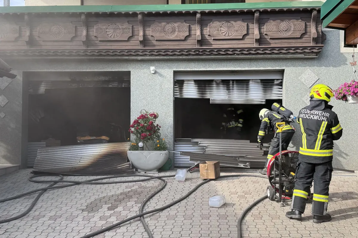 Drei Feuerwehrleute in Schutzkleidung arbeiten vor zwei verrauchten, geöffneten Garagentoren. Schläuche und Geräte sind auf dem Boden sichtbar.