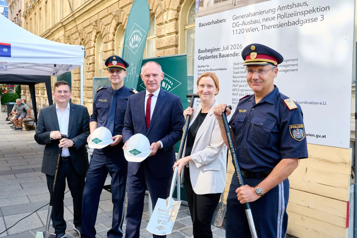 Mehrere Personen halten Spaten vor einem Poster, das ein Bauvorhaben in Wien beschreibt. Polizisten tragen Uniformen.