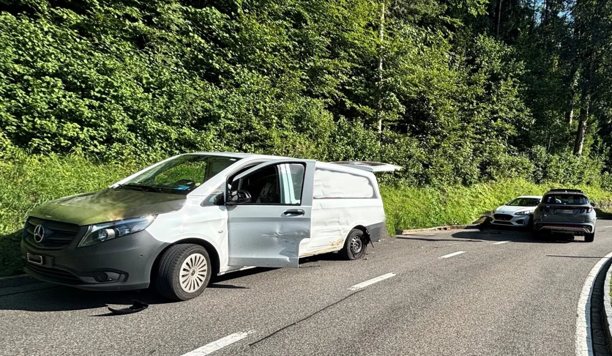 Ein weißer Mercedes-Transporter mit beschädigter Seite steht auf einer Straße am Waldrand. Zwei weitere Autos sind im Hintergrund zu sehen.