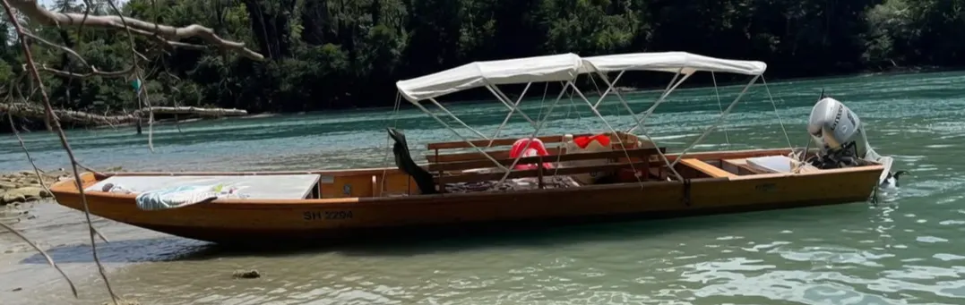 Ein hölzernes Boot mit weißem Sonnendach und Motor sitzt auf einem klaren Fluss. Dichte Waldvegetation säumt das Ufer im Hintergrund.