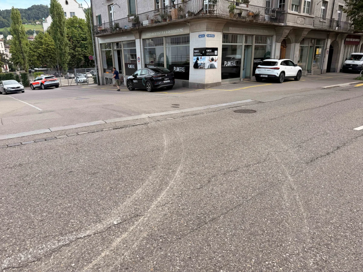 Eine Straßenecke mit mehreren geparkten Autos, darunter ein Polizeifahrzeug. Sichtbare Bremsspuren auf der Straße.