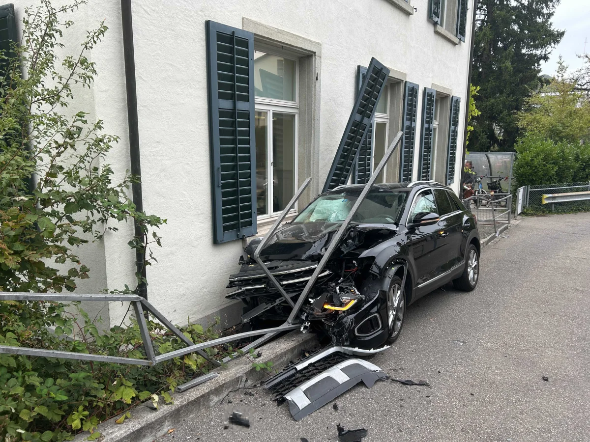Ein schwarzer SUV steht mit eingedrückter Front an der Wand eines Hauses, was zu sichtbaren Schäden geführt hat. Die Fensterläden sind aus ihrer Verankerung gerissen, und das Geländer ist verbogen.