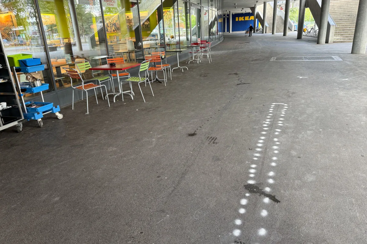 Ein leerer Gehweg mit weißen Sprühmarkierungen führt zu einem IKEA-Gebäude. Links stehen mehrere Tische und Stühle.