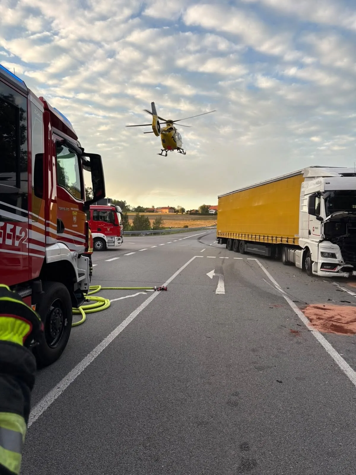 Ein Rettungshubschrauber fliegt über eine Unfallstelle, während Feuerwehrfahrzeuge und ein beschädigter Lkw zu sehen sind. Einsatzkräfte sind vor Ort und sichern die Stelle.