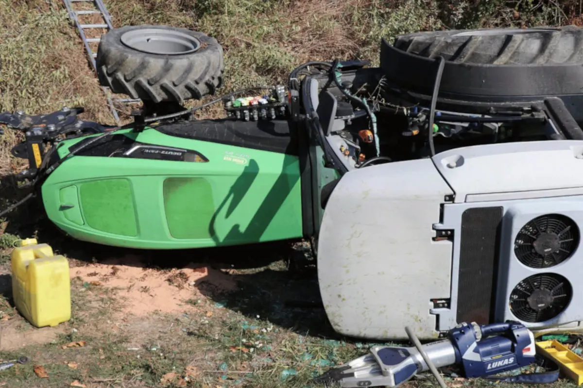 Ein grüner Traktor liegt umgekippt im Gras. Eine gelbe Kanne und Werkzeug liegen daneben.