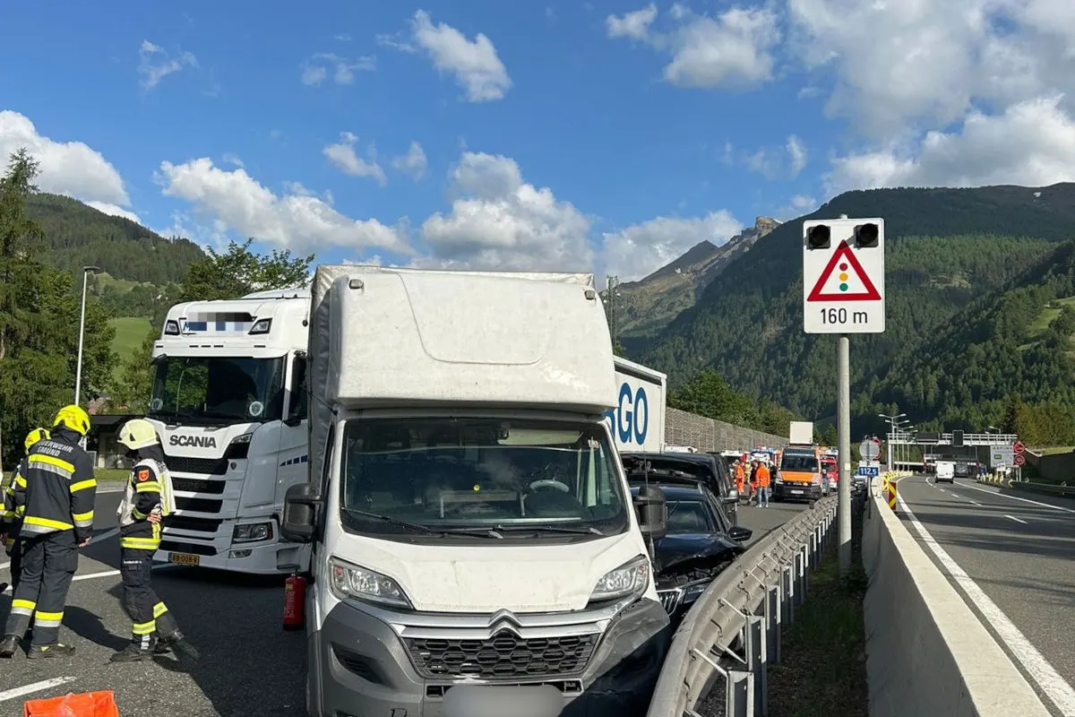Auf einer Autobahn sind ein Lkw und ein Transporter bei einem Unfall beteiligt, umgeben von Feuerwehrleuten in Einsatzkleidung. Im Hintergrund ist eine bergige Landschaft zu sehen.