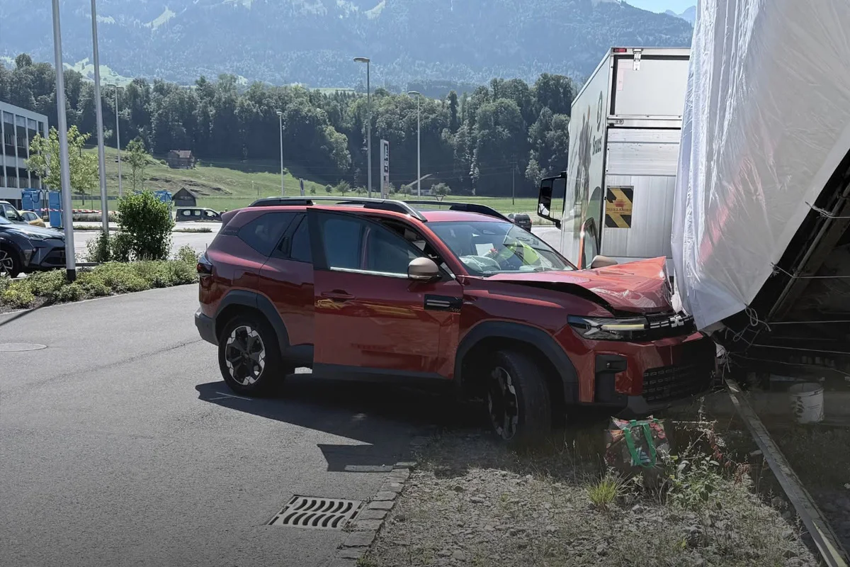 Ein roter SUV steht beschädigt auf einem Parkplatz, nachdem er frontal mit einem LKW-Anhänger kollidiert ist. Im Hintergrund ist eine ländliche Landschaft mit Bergen sichtbar.