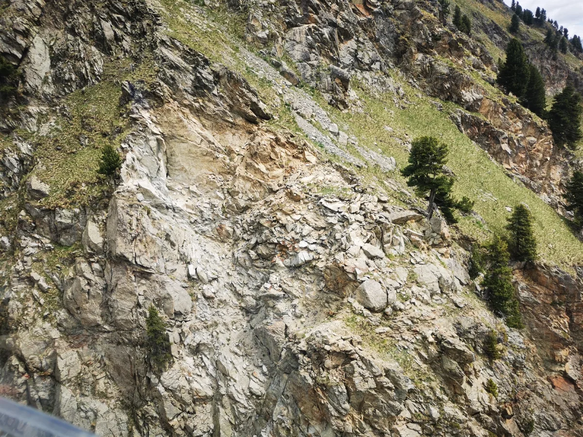Ein Berghang mit einem deutlichen Erdrutsch, der Teile eines Weges mit Felsen und Geröll überdeckt hat. Einige Bäume stehen auf der grünen Böschung.