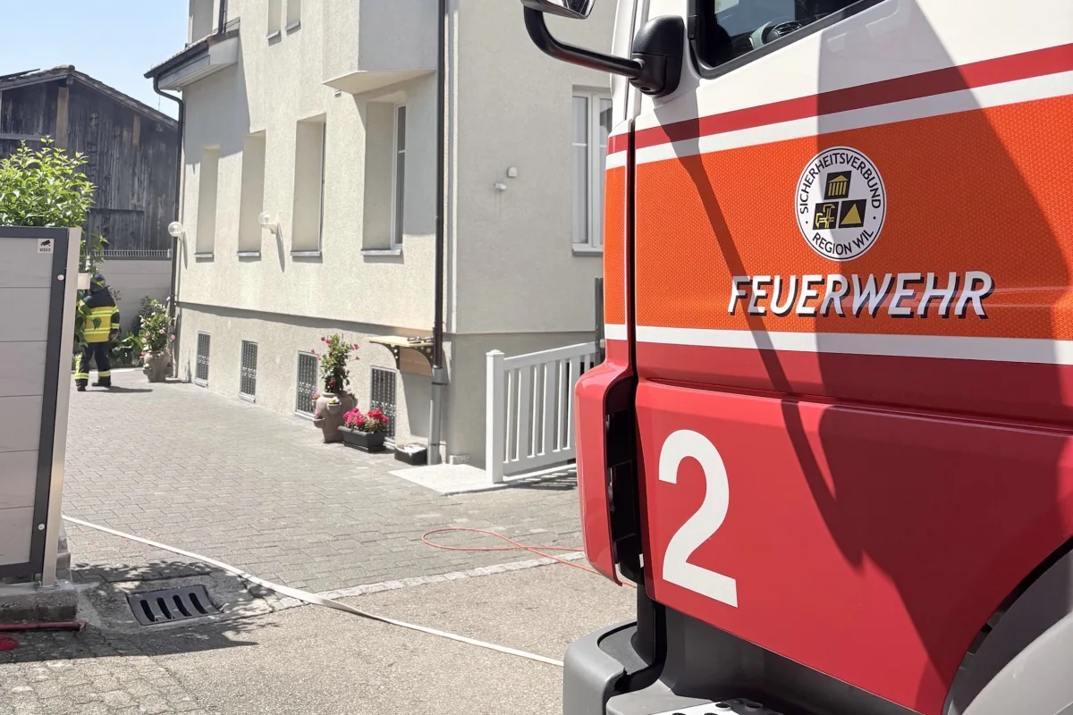 Ein rotes Feuerwehrfahrzeug mit der Aufschrift 'FEUERWEHR' steht vor einem Gebäude. Ein Feuerwehrmann geht in Richtung des Gebäudeeingangs.