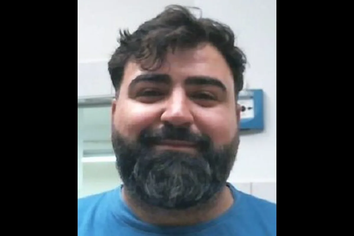 Ein lächelnder Mann mit vollem Bart blickt in die Kamera. Er trägt ein blaues T-Shirt und befindet sich in einem Innenraum.
