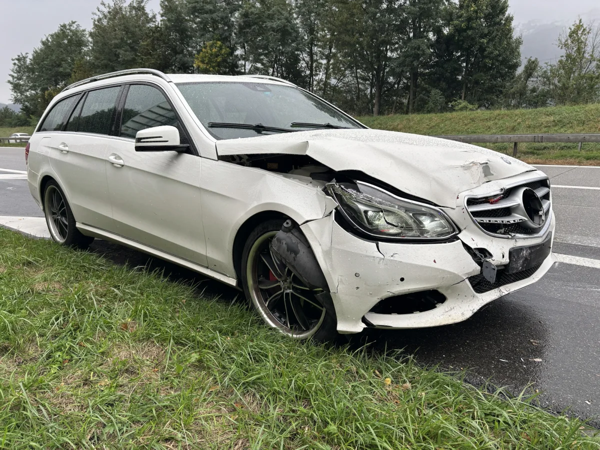 Ein weißer Mercedes mit schweren Frontschäden steht auf dem Seitenstreifen einer Straße. Die Stoßstange ist eingedrückt und Teile des Fahrzeugs fehlen.