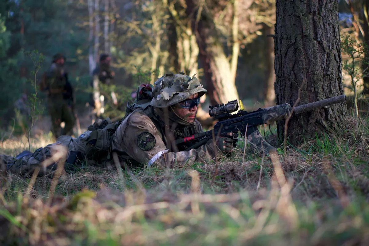 Ein Soldat in Tarnkleidung liegt mit einem Gewehr in einem Waldstück im Gras, während zwei weitere Soldaten im Hintergrund stehen.