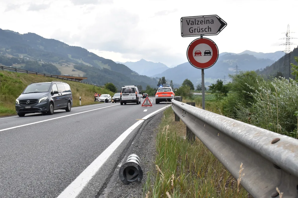 Auf einer Landstraße, gesäumt von Bergen, sichert ein Polizeiauto mit einem Warnschild eine Unfallstelle ab, während andere Fahrzeuge langsam passieren.