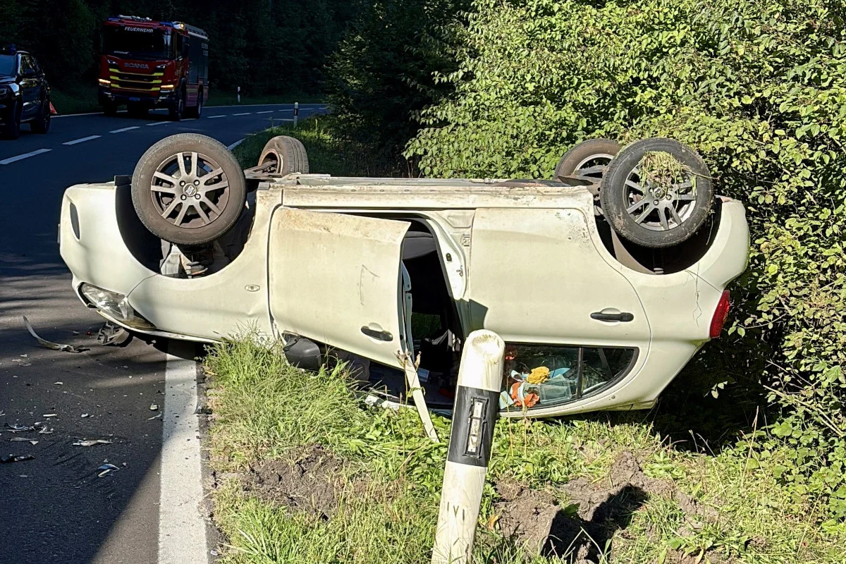 Ein beigefarbenes Auto liegt auf dem Dach neben einer Landstraße, mit sichtbaren Schäden und einem Feuerwehrauto im Hintergrund.