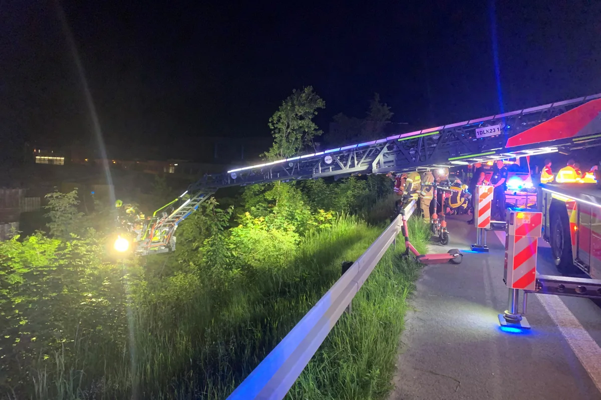 Bei Dunkelheit ist ein Feuerwehreinsatz zu sehen. Eine Drehleiter mit Scheinwerfer beleuchtet ein bewachsenes Gebiet, während mehrere Feuerwehrmänner anwesend sind.