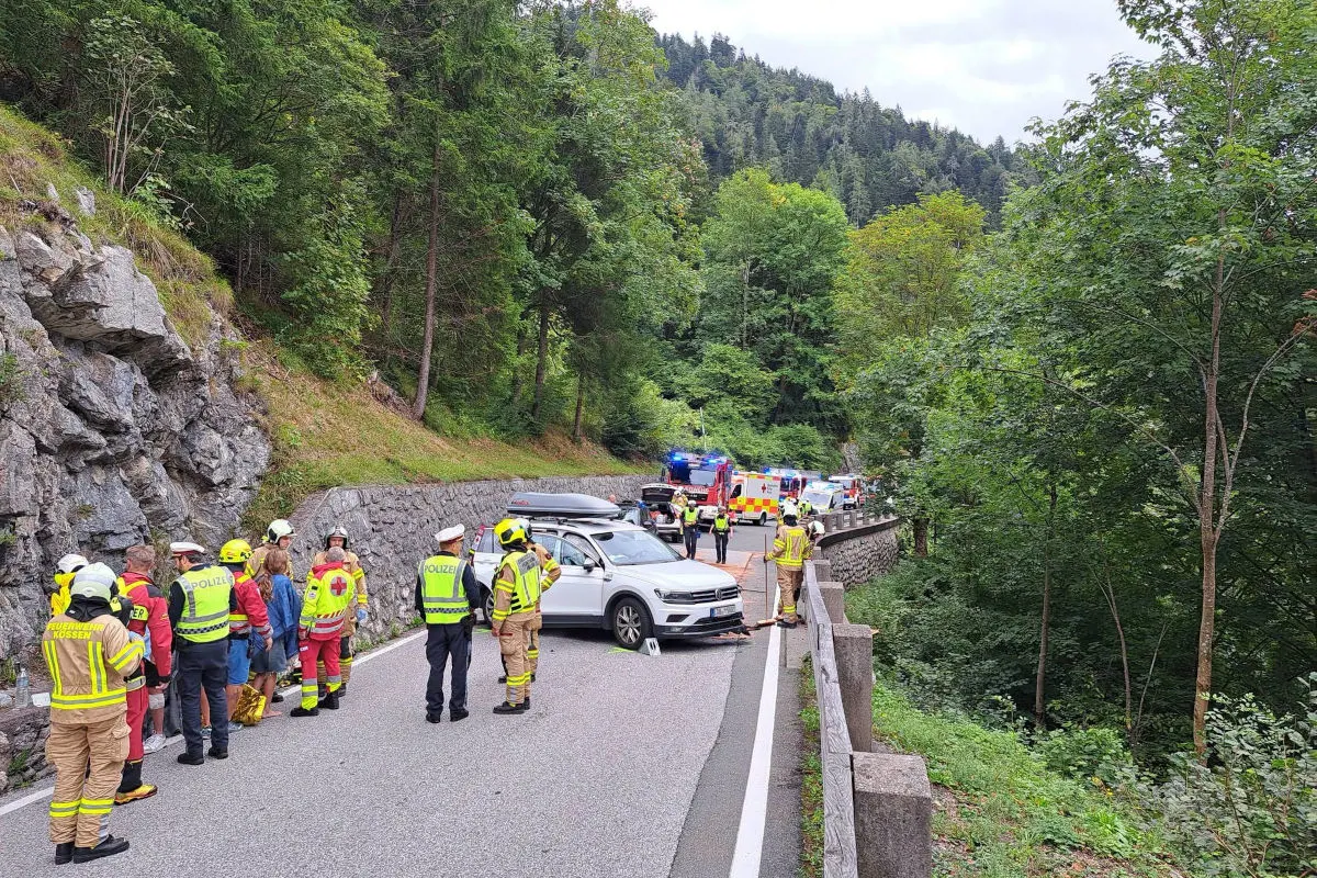 Mehrere Personen in Uniform und Rettungskräfte sind um ein am Straßenrand stehendem Auto versammelt. Im Hintergrund sind Polizei- und Feuerwehrfahrzeuge zu sehen.