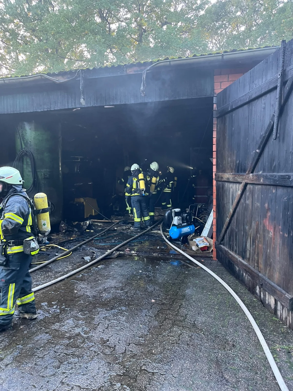 Mehrere Feuerwehrleute in voller Montur stehen vor dem Eingang einer verrauchten Lagerhalle. Schläuche und Ausrüstungsgegenstände sind im Vordergrund sichtbar.