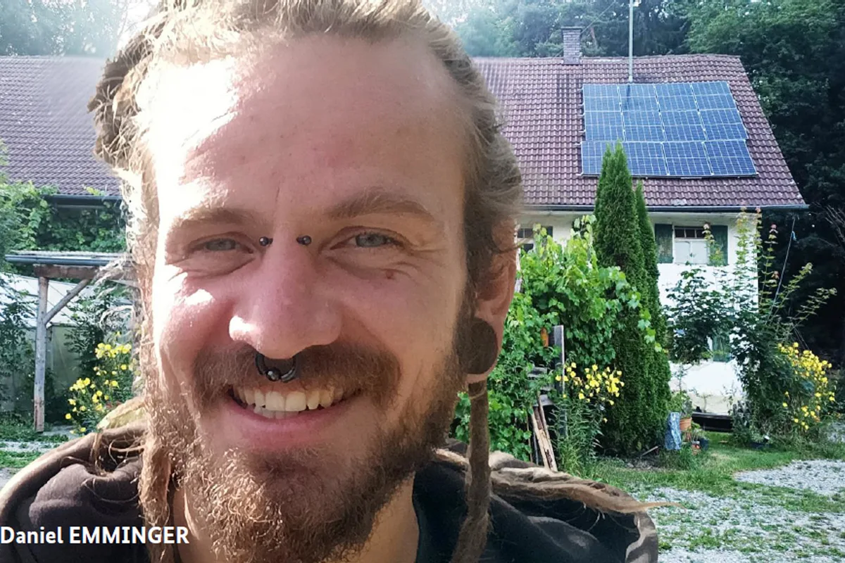 Ein Mann mit Piercings und Bart lächelt in die Kamera. Im Hintergrund ist ein Haus mit Solarpanelen auf dem Dach zu sehen.