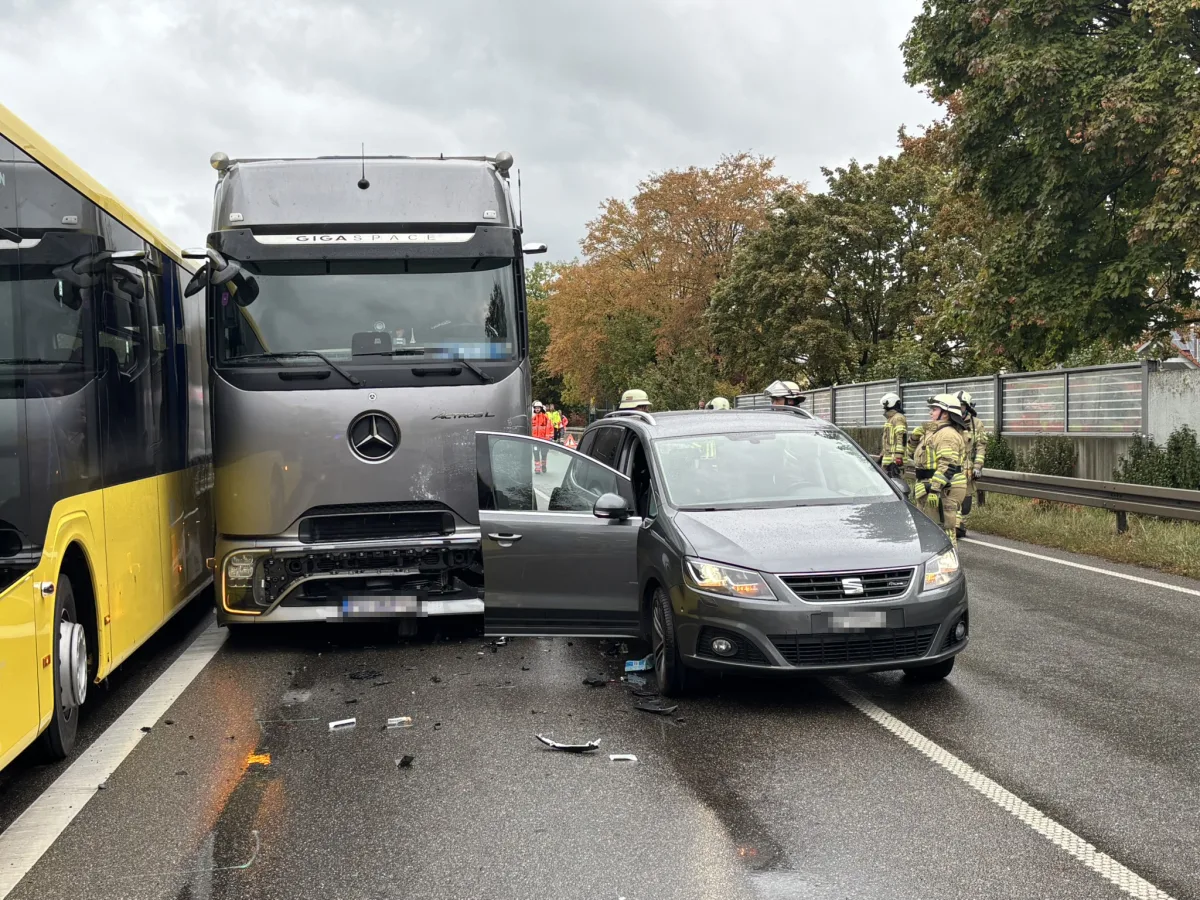 Ein grauer Lastwagen steht schräg zur Fahrbahn neben einem gelben Bus und einem grauen Pkw auf einer nassen Autobahn. Feuerwehrleute sind an der Unfallstelle.