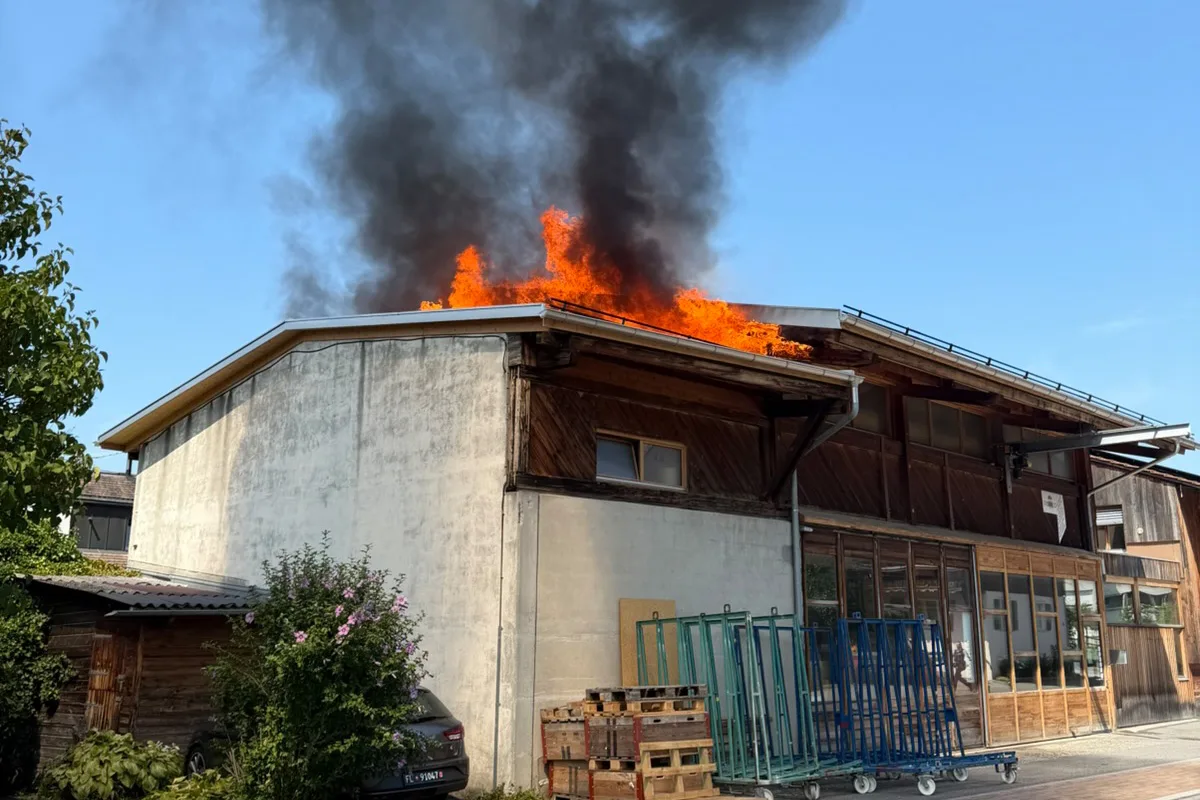 Flammen und schwarzer Rauch steigen vom Dach eines zweistöckigen Holzgebäudes auf. Im Vordergrund sind mehrere Paletten.