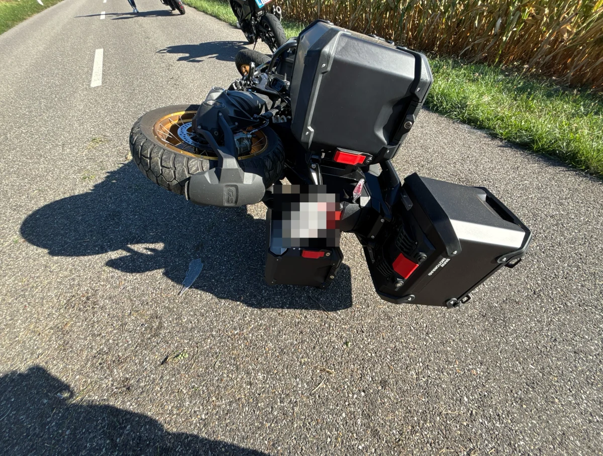 Ein Motorrad liegt umgestürzt auf einer asphaltierten Landstraße. Im Hintergrund sind weitere Motorräder zu erkennen.