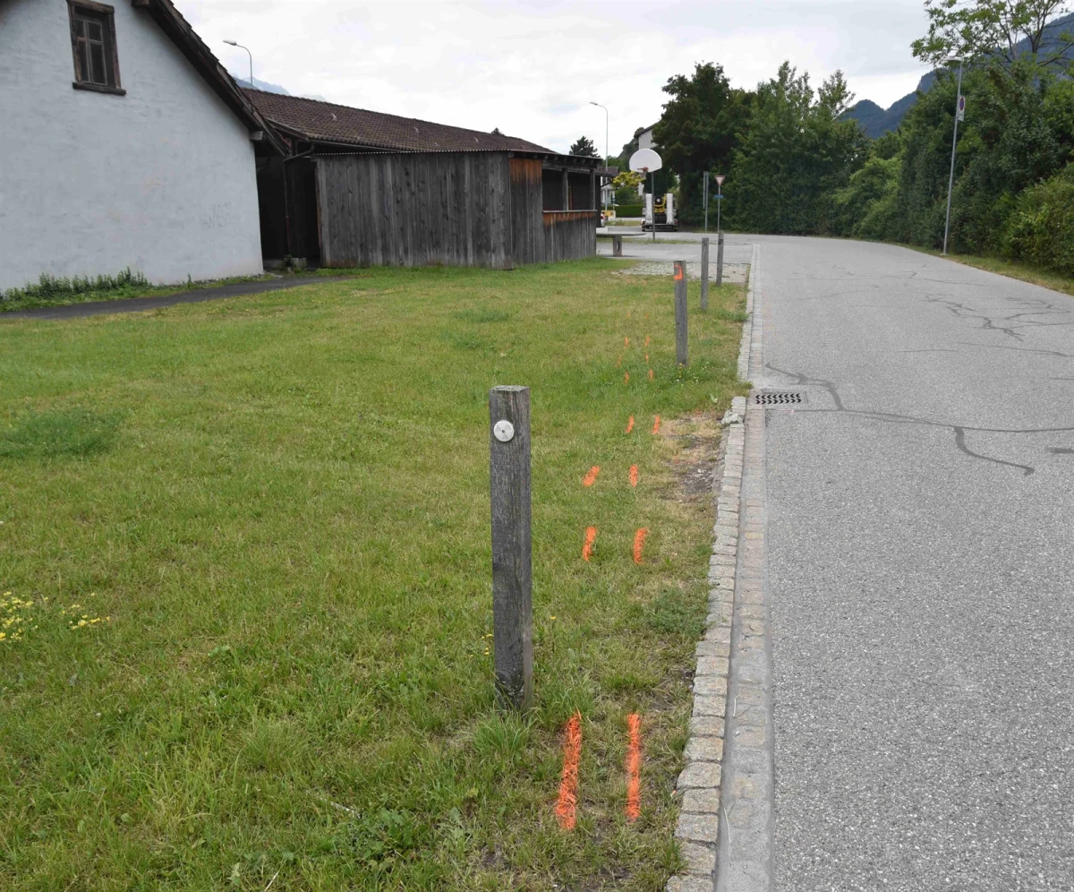 Zu sehen sind orange Markierungen auf dem Boden neben einer Straße, eingefasst von Holzpfosten und Gras.