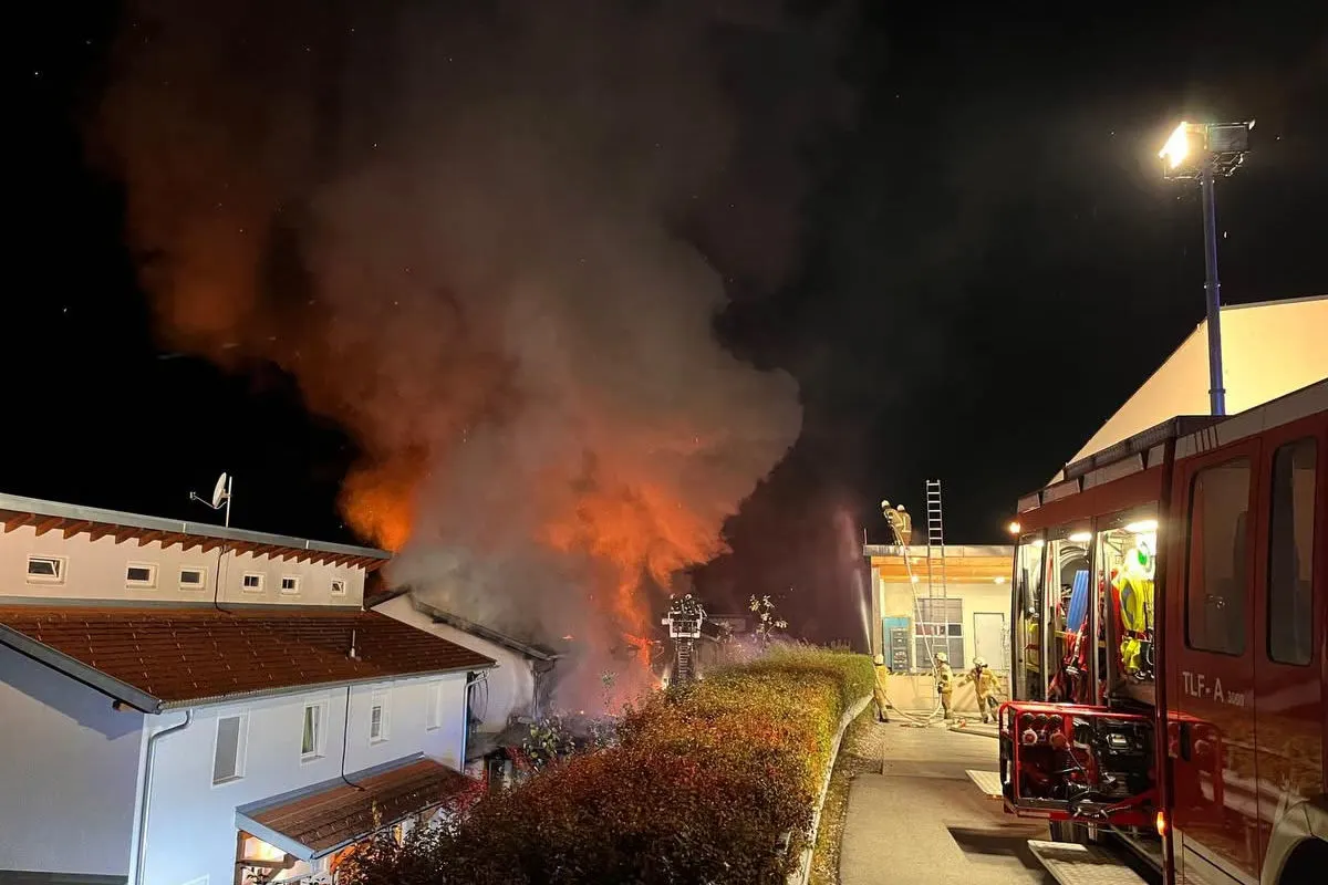 Feuerwehrkräfte bekämpfen einen Brand in einem Gebäude bei Nacht. Starker Rauch steigt auf, während ein Feuerwehrfahrzeug am Einsatzort steht.
