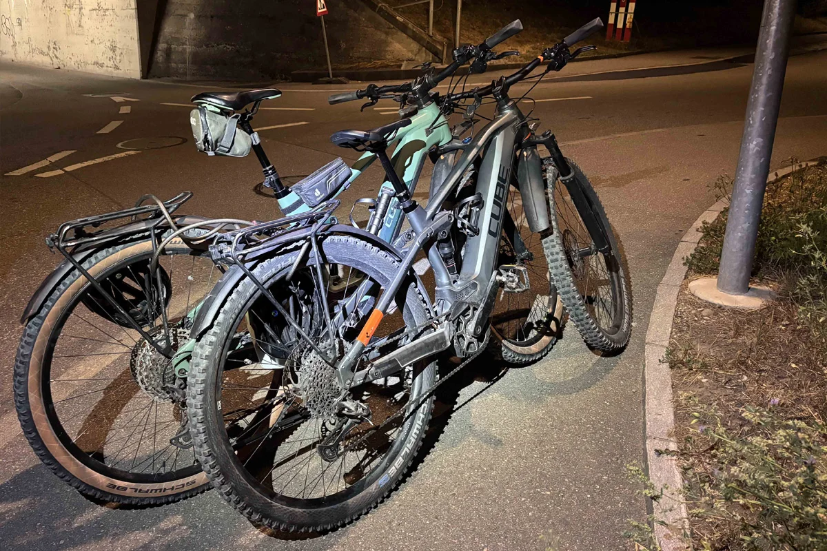 Zwei Mountainbikes stehen dicht nebeneinander auf einem Gehweg in der Nähe einer Unterführung. Die Szene ist bei Nacht aufgenommen und von einer Straßenlaterne beleuchtet.