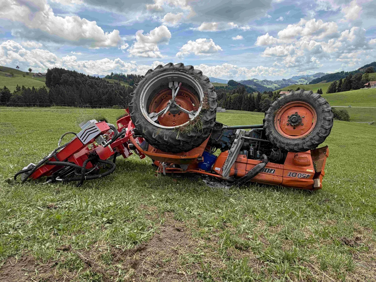 Ein umgekippter Traktor liegt auf einer grünen Wiese, umgeben von einer hügeligen Landschaft. Der Himmel ist mit Wolken bedeckt, und im Hintergrund sind Wälder und Felder sichtbar.