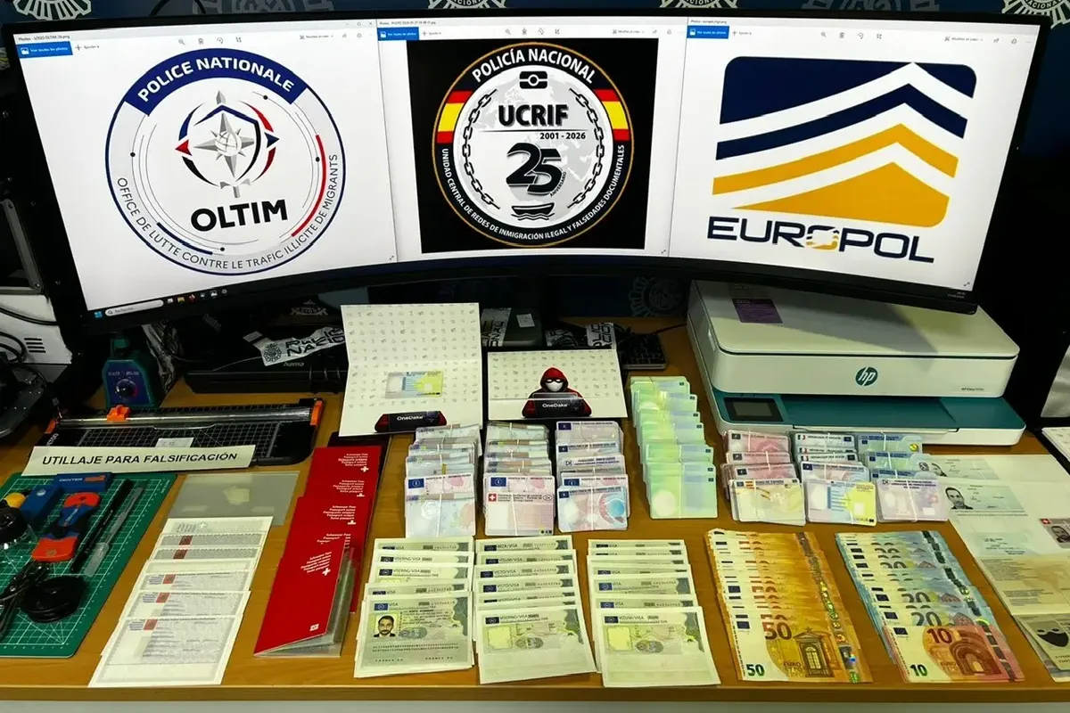 Auf einem Tisch sind gefälschte Ausweise, Geldscheine und Geräte zur Fälschung ausgebreitet. Im Hintergrund sind drei Bildschirme mit Logos von Polizeibehörden zu sehen.