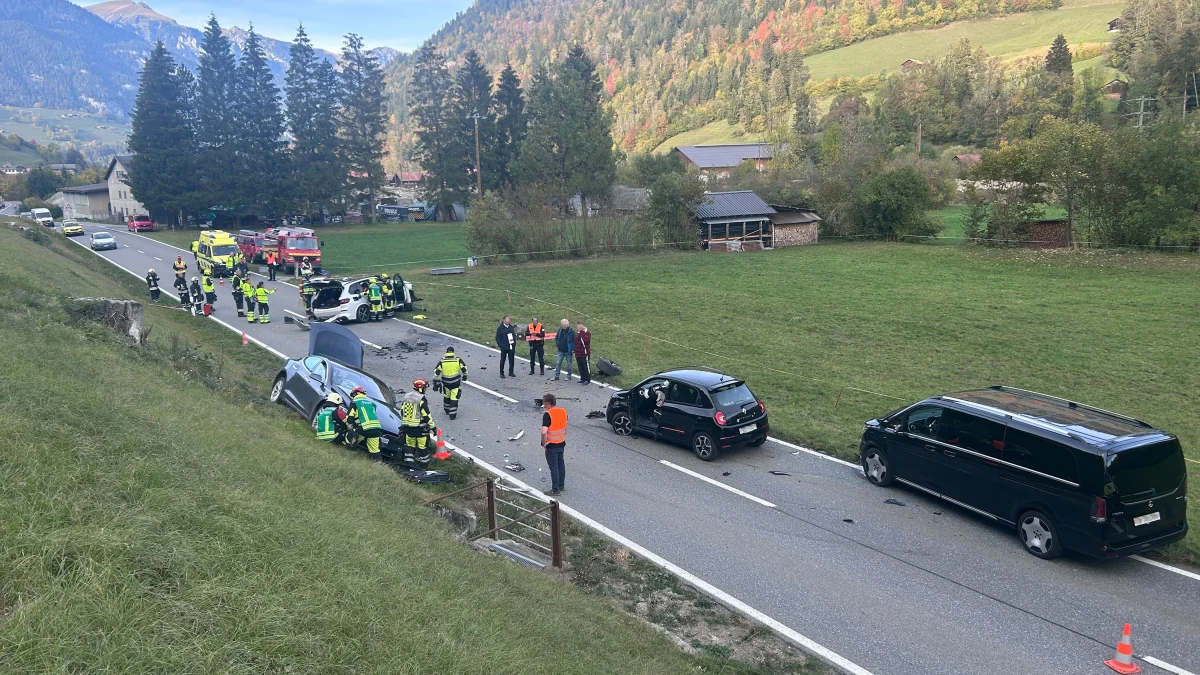 Auf einer Landstraße sind mehrere verunfallte Fahrzeuge zu sehen, umgeben von Einsatzkräften der Feuerwehr. Einige Menschen stehen daneben.