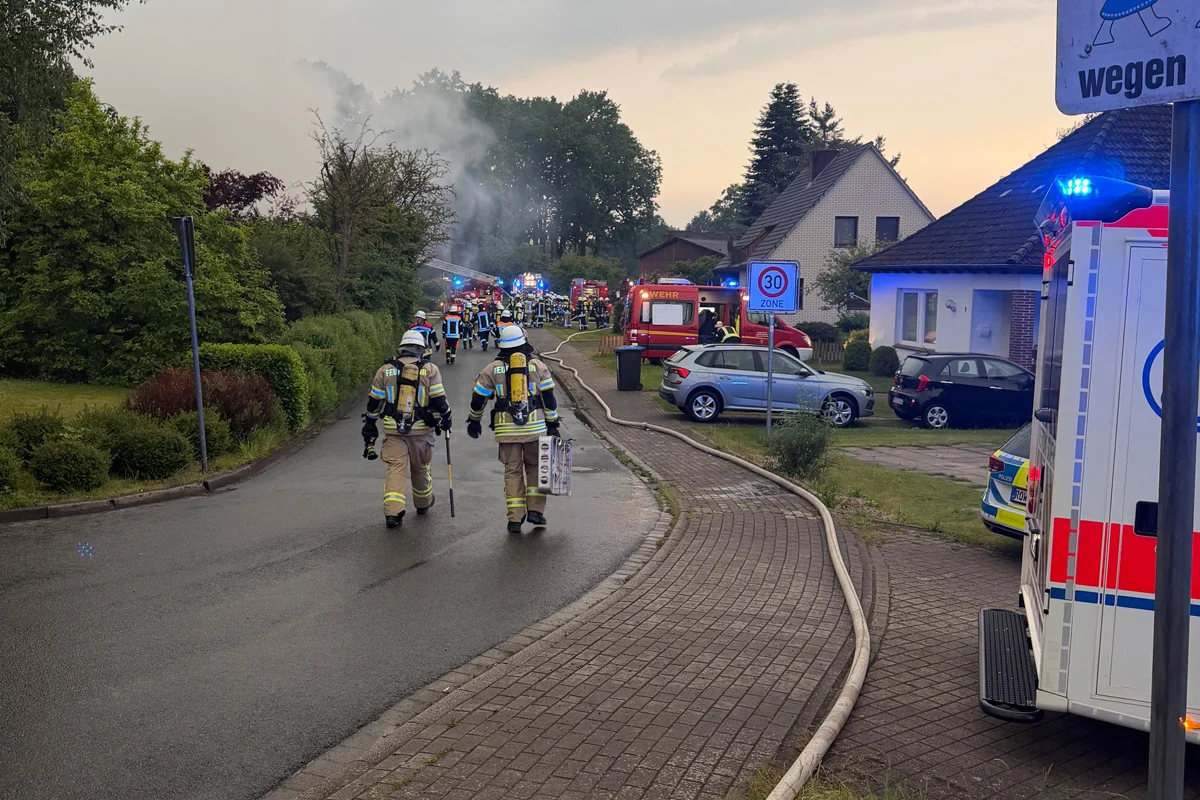 Mehrere Feuerwehrleute betreten eine Wohnstraße, deren Ende von Rauch verstellt ist. Zahlreiche Einsatzfahrzeuge sind sichtbar.