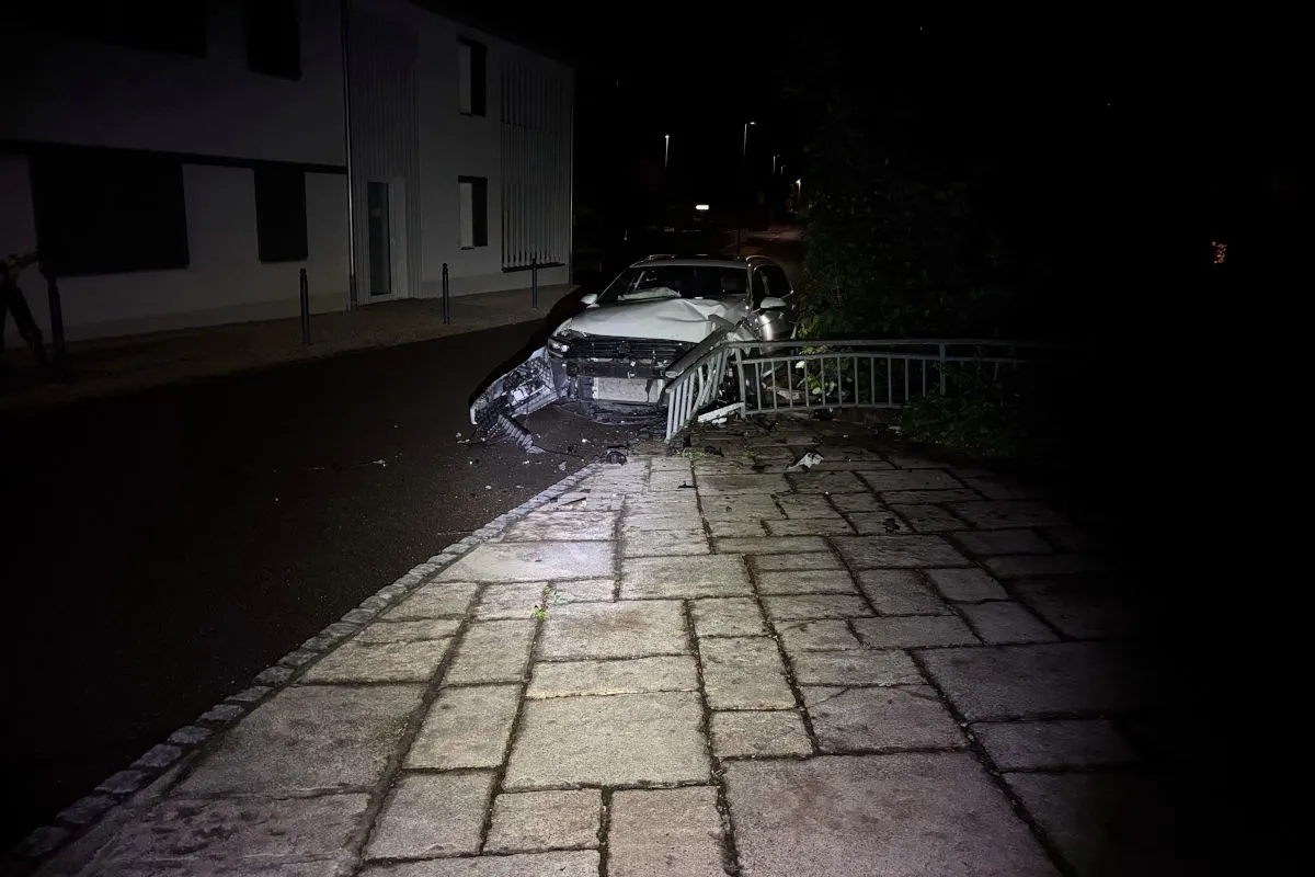 Ein silbernes Auto steht stark beschädigt frontal an einem Metallgeländer auf einem Gehweg. Die Szene ist nachts beleuchtet.