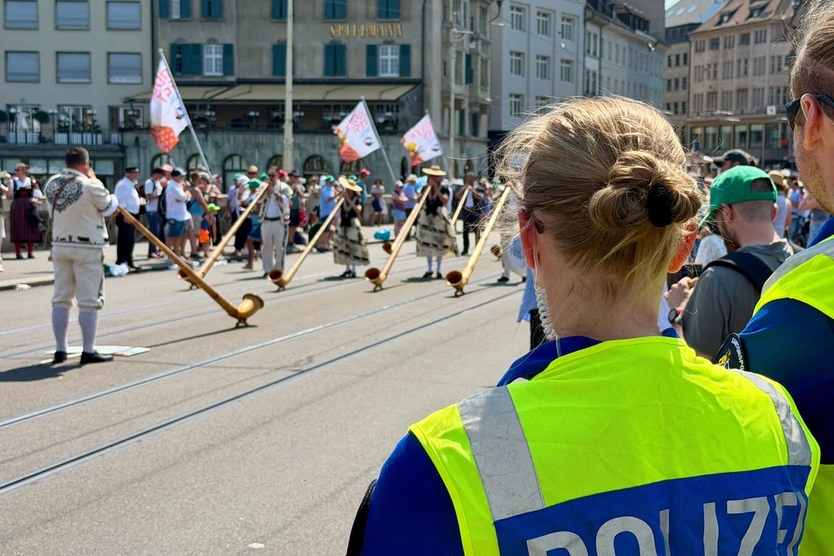 Eine Polizistin, von hinten zu sehen, beobachtet eine Gruppe von Alphornspielern auf einer Straße. Zuschauer stehen im Hintergrund.