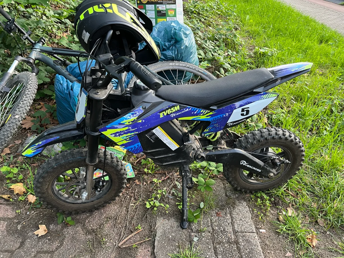 Ein kleines, buntes Motorrad mit der Aufschrift 'EV65M' steht auf einem Weg neben einem Fahrrad und einem Helm.