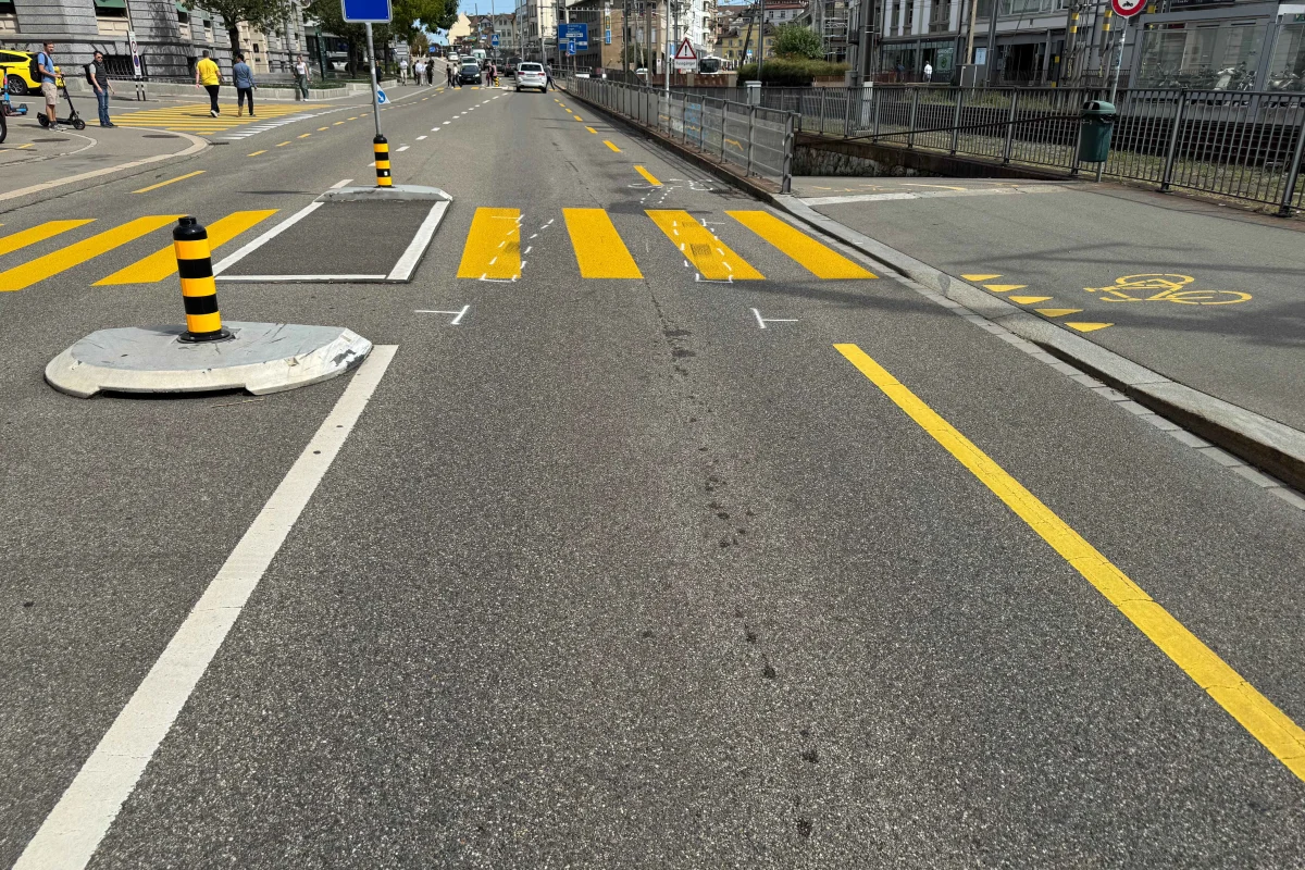 Eine städtische Straße mit einem neu markierten Zebrastreifen und zwei Pollern. Fuß- und Radwege sind sichtbar, Personen im Hintergrund.