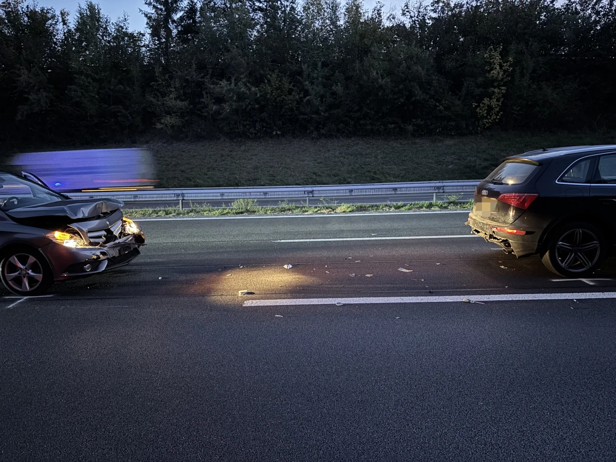 Zwei Autos mit sichtbaren Front- und Heckschäden stehen auf einer Autobahn. Es ist dämmerig und ein Fahrzeug mit Blaulicht fährt im Hintergrund vorbei.