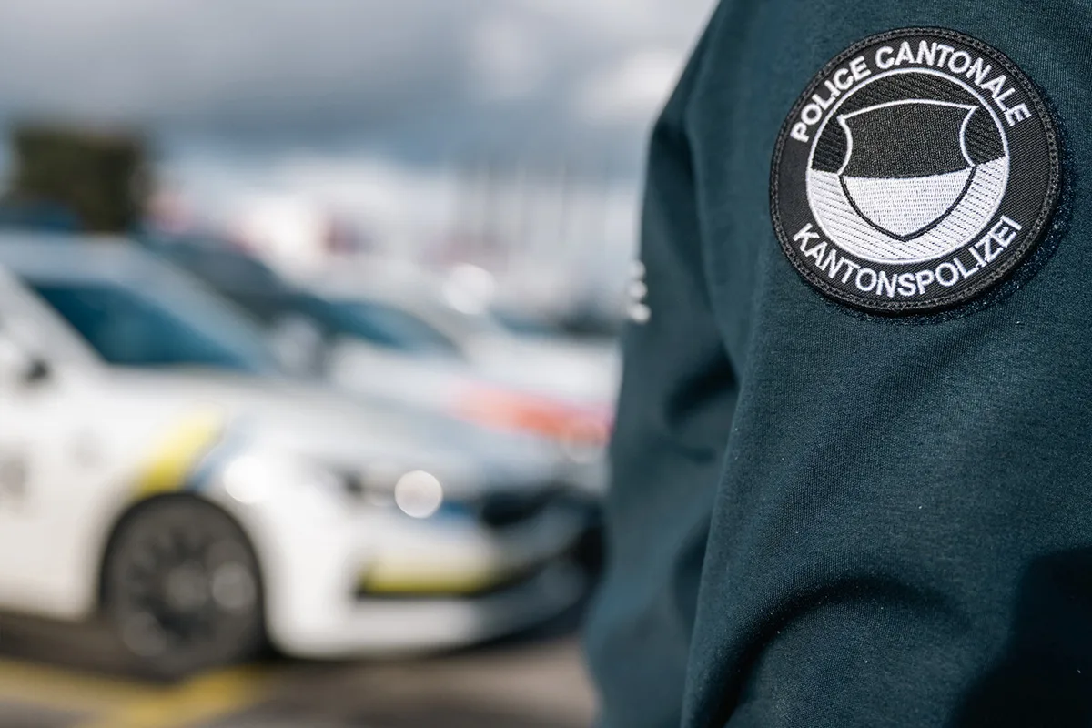 Das Bild zeigt ein Abzeichen der Kantonspolizei auf der Uniform eines Polizeibeamten, im Hintergrund unscharf ein Polizeifahrzeug.
