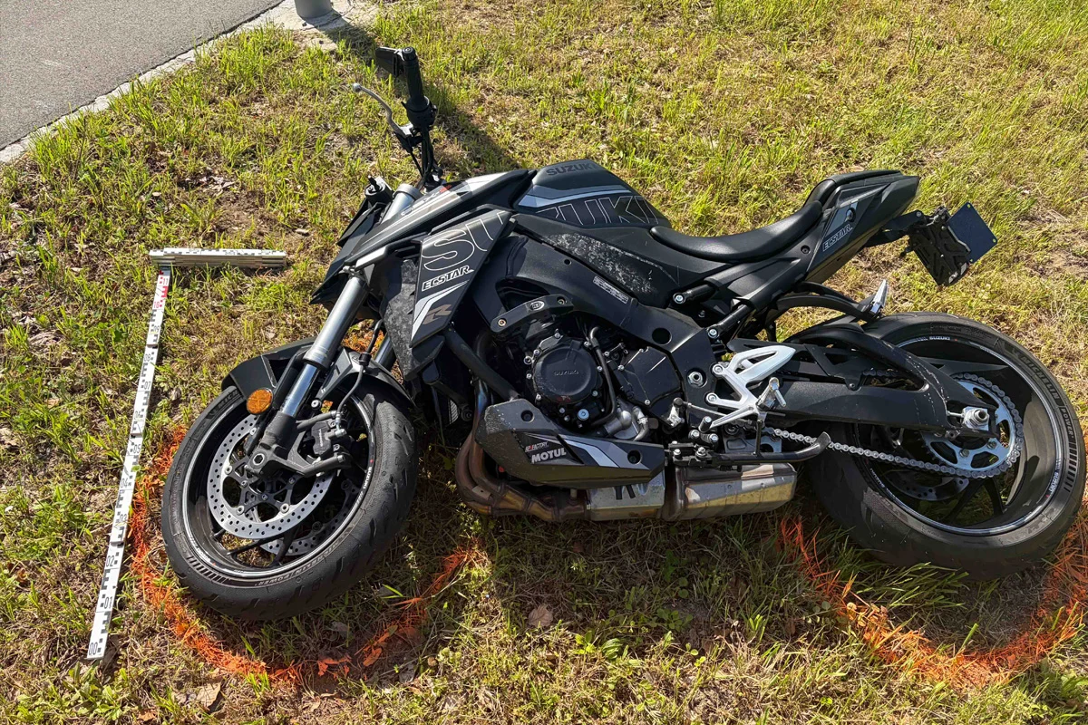 Ein schwarzes Motorrad liegt auf der Seite in einer Wiese neben einer Straße. Es sind orange Markierungen und ein Messstab sichtbar.