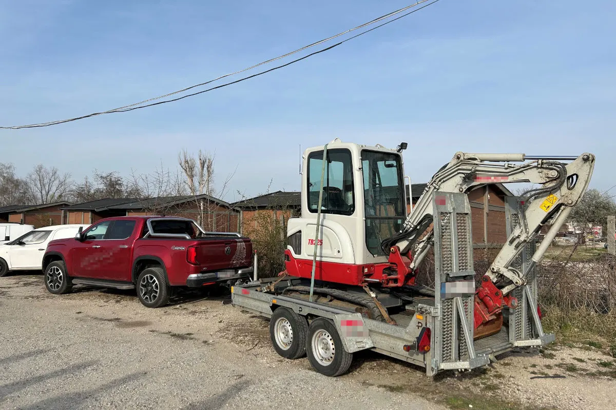 Ein roter Pickup-LKW steht neben einem Anhänger, auf dem ein Bagger transportiert wird, in einem ländlichen Wohngebiet.