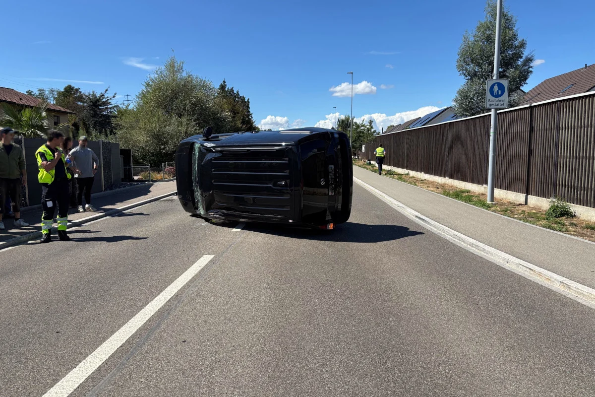 Ein schwarzes Auto liegt seitlich auf einer Straße. Menschen stehen in der Nähe und ein Polizist ist zu sehen. Es ist ein sonniger Tag.