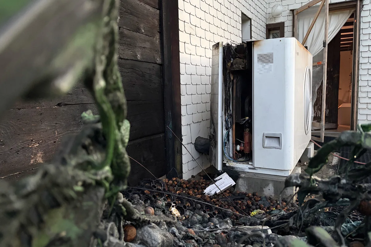 Eine beschädigte und verkohlte Wärmepumpe steht an einer Gebäudeaußenwand, wo Spuren eines Brandes sichtbar sind. Eine benachbarte Tür ist offen, und das Innenleben der Pumpe ist freigelegt.