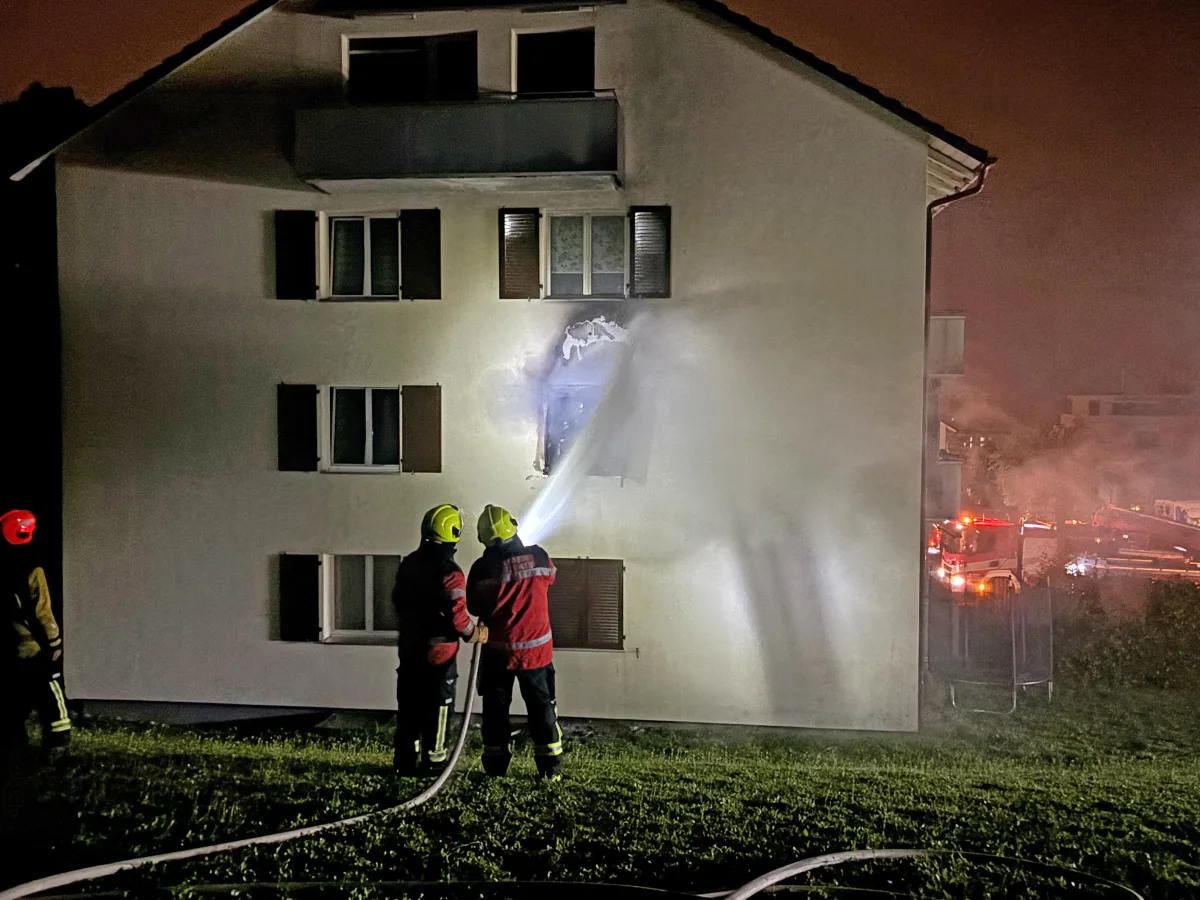 Mehrere Feuerwehrleute bekämpfen einen Brand an einem Wohnhausfenster bei Nacht. Eindämmung des Feuers mit Löschwasser.