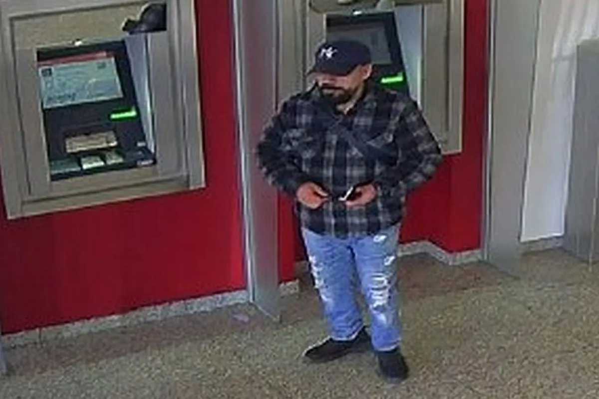 Ein Mann steht in einer Bankfiliale vor einem Geldautomaten. Er trägt eine karierte Jacke, eine Baseballmütze und hält ein Mobiltelefon in der Hand.