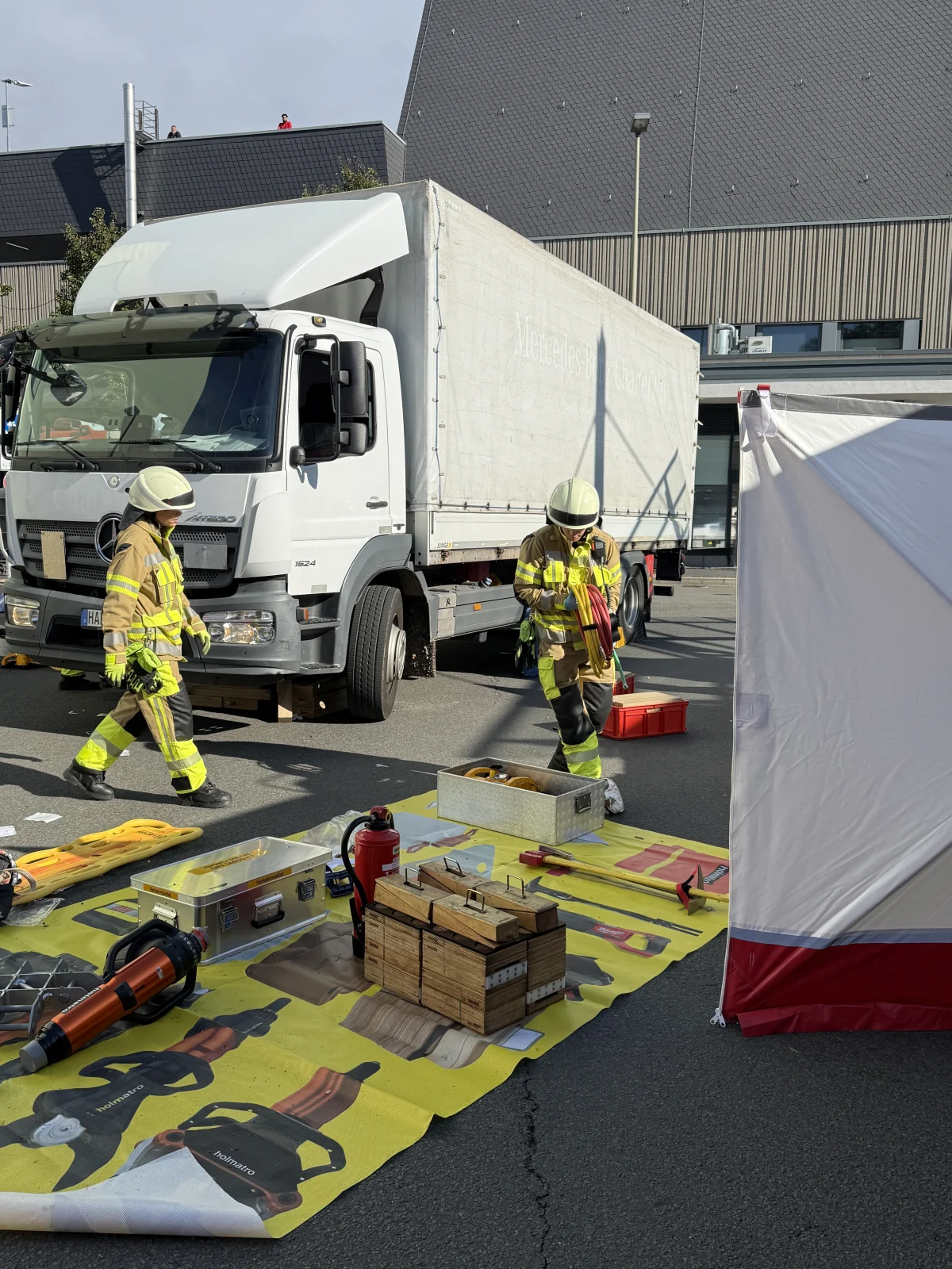 Zwei Feuerwehrleute in Schutzkleidung stehen vor einem weißem Lastwagen mit offener Fahrerkabine und vorbereiteter Ausrüstung auf dem Boden.